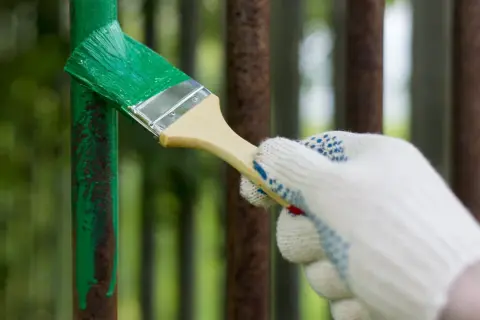 Mano guantata che inizia a verniciare un cancello in ferro con vernice verde brillante, coprendo la ruggine.