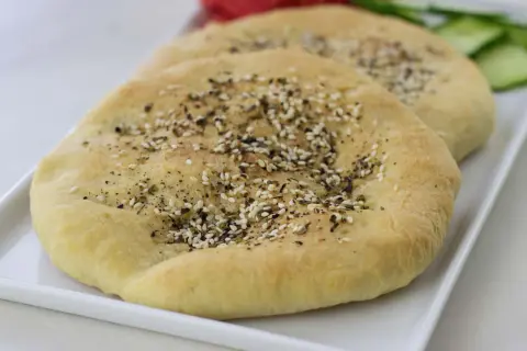 Två runda manakish zaatar med sesamfrön och örter på en vit bricka, med skivad gurka och tomat i bakgrunden.