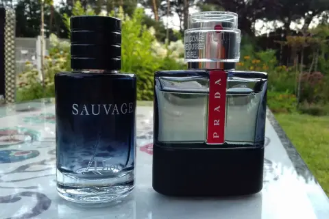 Dwie butelki perfum: Dior Sauvage i Prada Carbon, stoją na stoliku w ogrodzie.