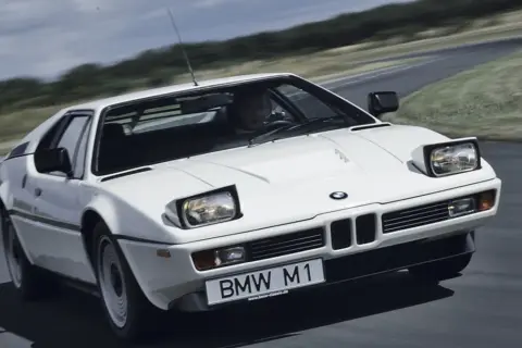 Białe BMW M1 na zakręcie drogi, z widocznym kierowcą i tablicą rejestracyjną.