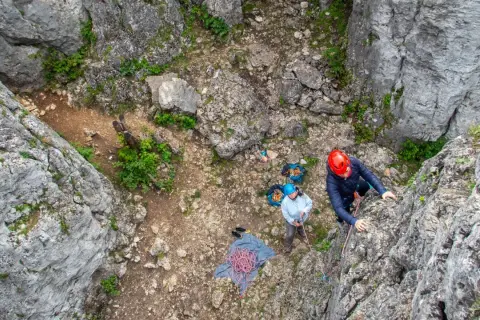 Wspinaczka po skale. Dwie osoby w kaskach przygotowują się do wspinaczki, liny i sprzęt leżą na ziemi.