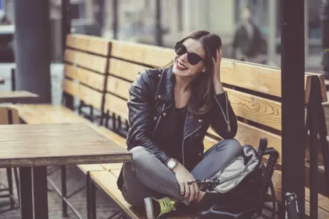 Dziewczyna w skórzanej kurtce i okularach przeciwsłonecznych, z uśmiechem na twarzy, siedzi na ławce w kawiarni. Jej styl jest zdecydowanie rockowy.