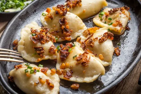 Pyszne pierogi z mięsem, posypane skwarkami i szczypiorkiem, podane na żeliwnej patelni.