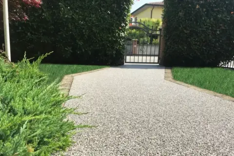 Vialetto carrabile in pietra, con ghiaia chiara e bordi in cotto, conduce a un cancello in ferro battuto.