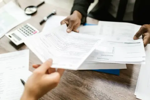 Dwie osoby analizują dokumenty. Na stole leży kalkulator i lupa. Widać fragmenty formularzy, jeden z nich jest podawany.