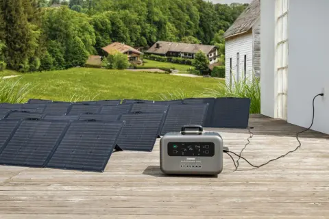 Instalación de baterías solares en un porche de madera, con paneles solares desplegados y una unidad de energía portátil conectada a una casa.