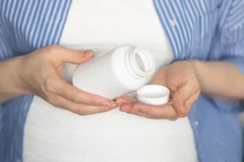 Kobieta w ciąży trzyma białą buteleczkę i jej nakrętkę. Rozważa, czy wziąć paracetamol w ciąży.