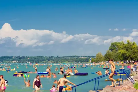 Tłum ludzi cieszy się słońcem i wodą na zatłoczonej balaton plaży. Wiele osób pływa, relaksuje się na materacach i bawi się w wodzie.