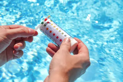 Manos sostienen una tira reactiva para medir el pH de la piscina. El agua azul y cristalina de la piscina se ve al fondo.