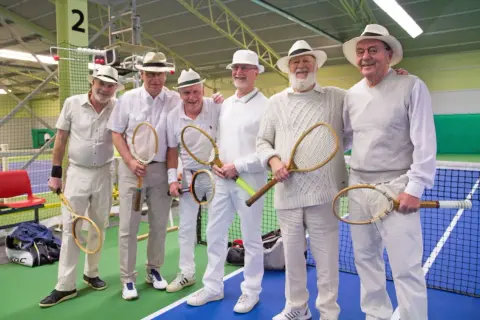 Sex äldre herrar i hattar och vita kläder poserar med tennisracketar, redo för en match i stil med Gustaf V:s tennis.