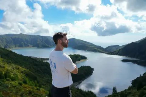 Hombre contemplando la **Lagoa do Fogo**, un lago azul rodeado de montañas verdes y un cielo nublado.