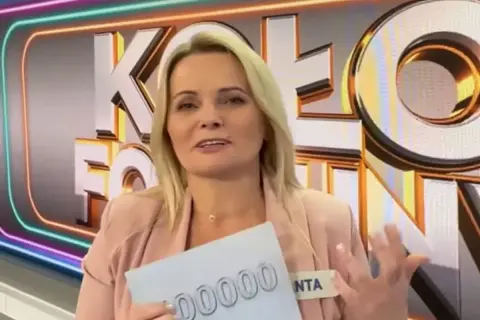 Kobieta w różowym żakiecie trzyma kartkę z napisem "00000" na tle logo "Koło Fortuny".