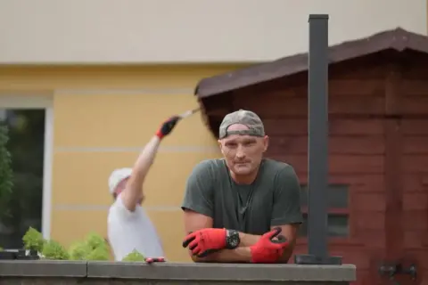 Mężczyzna w czapce i rękawiczkach opiera się o mur. W tle inny mężczyzna maluje drewnianą szopę. Jak zgłosić się do polowania na ogród?