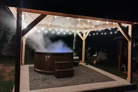 Drewniana beczka z gorącą wodą, z której unosi się para, pod zadaszeniem ozdobionym lampkami. Idealne miejsce na relaks w stylu sauna fińska.