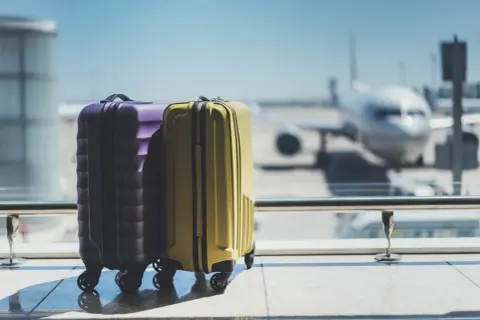 Zwei Koffer, lila und gelb, stehen am Flughafen vor einem Flugzeug.