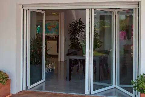 Puertas de cristales plegables que abren a un comedor interior con plantas y una mesa.