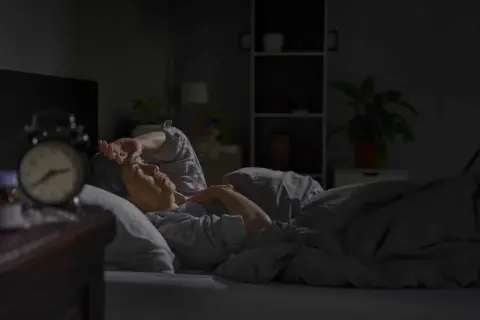 A person lies awake in bed, hand on forehead, experiencing insomnia after surgery. An alarm clock sits on the nightstand.
