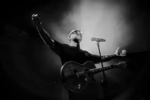 Artysta z gitarą na scenie, w okularach, z podniesionymi rękami.