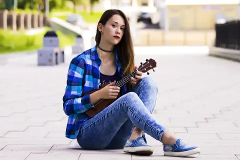 Dziewczyna w kratę i jeansach pokazuje, jak trzymać ukulele, siedząc na chodniku.