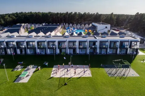Widok z lotu ptaka na kompleks domków i budynków mieszkalnych z basenem i placem zabaw, na trawniku boisko do siatkówki i stoły do ping-ponga.