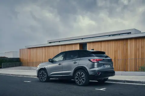 Vue arrière d'un SUV gris Renault Austral, stationné sur une route devant un bâtiment moderne en bois. La motorisation est performante.