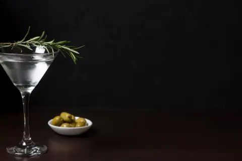Elegancki drink z oliwką i gałązką rozmarynu, obok miseczka z zielonymi oliwkami.
