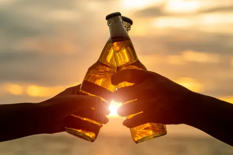 Dwie ręce wznoszą butelki piwa do toastu o zachodzie słońca. W Niemczech od ilu lat można pić alkohol? Od 16 roku życia.