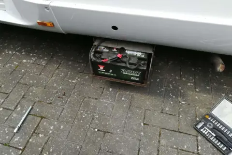 Emplacement batterie Ford Transit : une batterie Yuasa "Active Leisure & Marine" est visible sous le véhicule, dans un support métallique. Une clé à douille et une boîte à outils sont au sol.