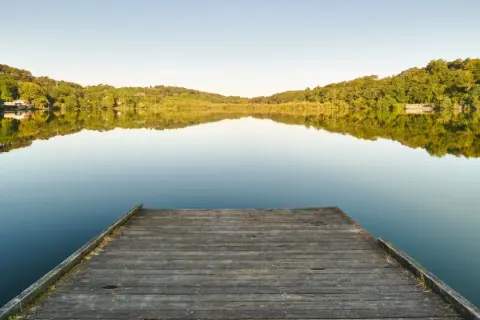 Un ponton en bois mène au lac Marion de Biarritz, reflétant les collines verdoyantes sous un ciel clair.
