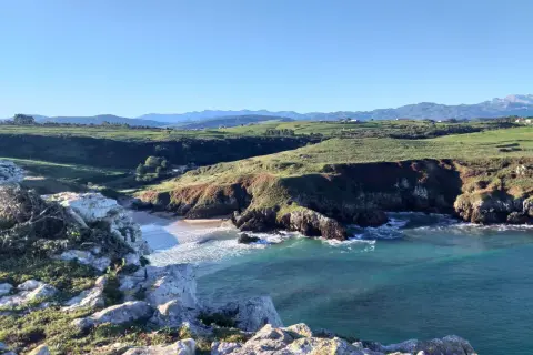 Acantilados y mar turquesa, ideales para rutas senderismo San Vicente de la Barquera. Paisaje costero con verdes colinas y montañas al fondo.