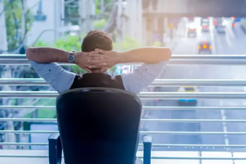 Uomo con le mani dietro la testa, seduto su una sedia, guarda il traffico. Forse è un momento di **riposo compensativo non goduto**.