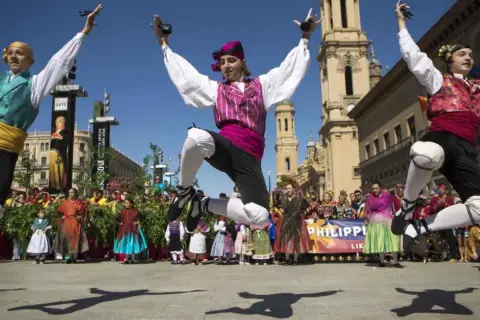 Jóvenes bailan jotas del Pilar con castañuelas, saltando enérgicamente. Al fondo, gente vestida de época y edificios históricos.