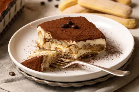 Ein Stück Tiramisu, zubereitet nach einem einfachen tiramisu rezept, liegt auf einem Teller. Kakaopulver bestäubt die cremige Schicht und die Löffelbiskuits.
