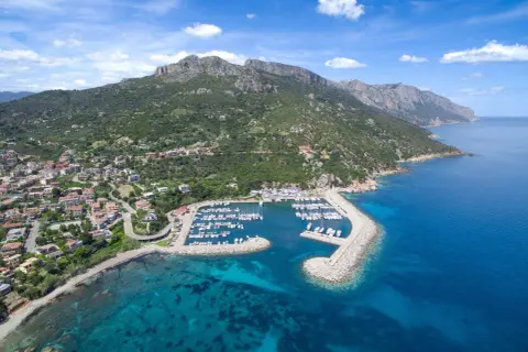 Baunei: un porto turistico con barche ormeggiate, case colorate e montagne verdi che si tuffano nel mare cristallino. Le spiagge vicine invitano al relax.