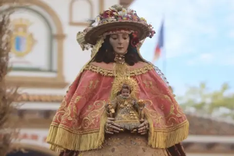 Virgen del Rocío pastora, con su manto rosa bordado y el Niño Jesús en brazos, adornada con flores y un sombrero.