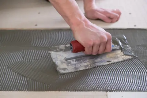A hand uses a notched trowel to spread thinset for cement board, creating a textured surface for tile adhesion.