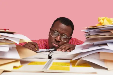 Un homme fatigué, entouré de piles de documents, semble avoir un avis de départ. Il repose sa tête sur un livre ouvert, l'air dépassé par la paperasse.