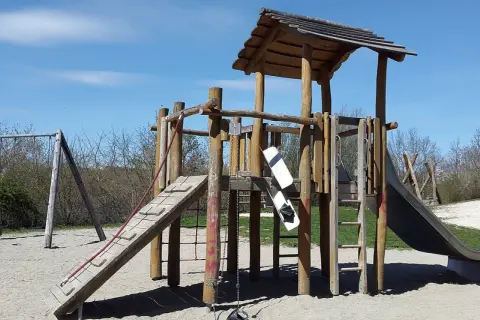 Drewniany plac zabaw ze zjeżdżalnią, drabinką i huśtawką na piaszczystym podłożu.