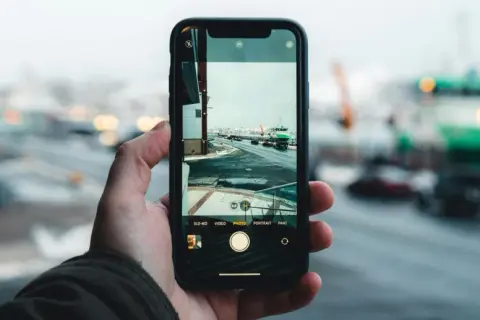 Dłoń trzyma smartfon, na ekranie którego widać interfejs aparatu. Pokazuje, jak dziala aparat, rejestrując zimowy krajobraz z drogą i zielonym statkiem.