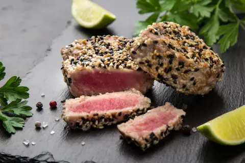 Apetyczny stek z tuńczyka w sezamie, z kawałkami limonki i natką pietruszki na czarnym kamieniu.