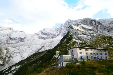 Un rifugio in pietra con persiane blu si staglia su un pendio verdeggiante, sotto imponenti montagne rocciose. Un esempio di rifugi in Veneto.