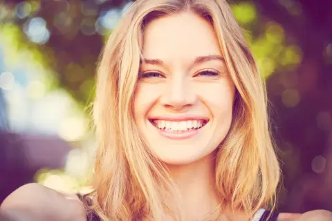 Strahlendes Lächeln einer jungen Frau mit blonden Haaren, die eine natürliche Frisur trägt.