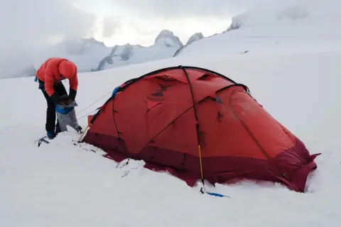 Osoba w pomarańczowej kurtce przygotowuje sprzęt przy czerwonym namiocie. W tle majestatyczne, ośnieżone szczyty, zapowiadające trudne wejście na Mount Everest.