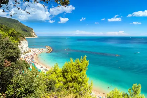 Spiaggia affollata con ombrelloni colorati e gente che nuota nelle acque cristalline. Una scogliera bianca domina la costa, incorniciata dal verde della vegetazione. Una perfetta località mare Marche.