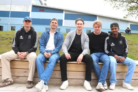 Fem unga män, potentiella k1 fighters, sitter på en träbänk framför en blå och grå byggnad.