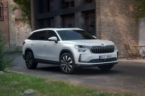 Biały Skoda Kodiaq 2025 zaparkowany przed ceglanym budynkiem, z zielenią i stolikiem z krzesłami w tle.