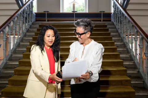 Två kvinnor diskuterar papper på en trappa. Den ena ger den andra en telefon, kanske för att hantera administrativa uppgifter.
