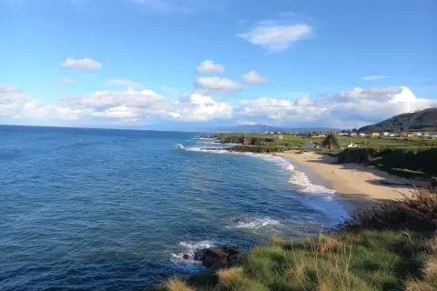 Mar azul y cielo nublado sobre la playa de Xuncos, con casas dispersas en la costa verde.