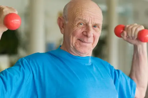 Ein lächelnder älterer Mann macht Muskeltraining im Alter mit roten Hanteln.