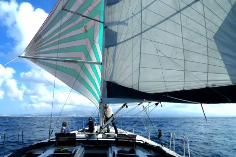 Vela colorata e bianca gonfie al vento, con persone a bordo pronte a mettersi alla cappa. Mare blu e cielo con nuvole bianche.